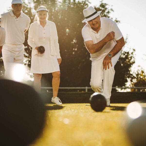 Senior people playing boules in a park Senior man in hat bending forward to throw a boules in a lawn. Elderly man playing boules in a lawn with his friends.