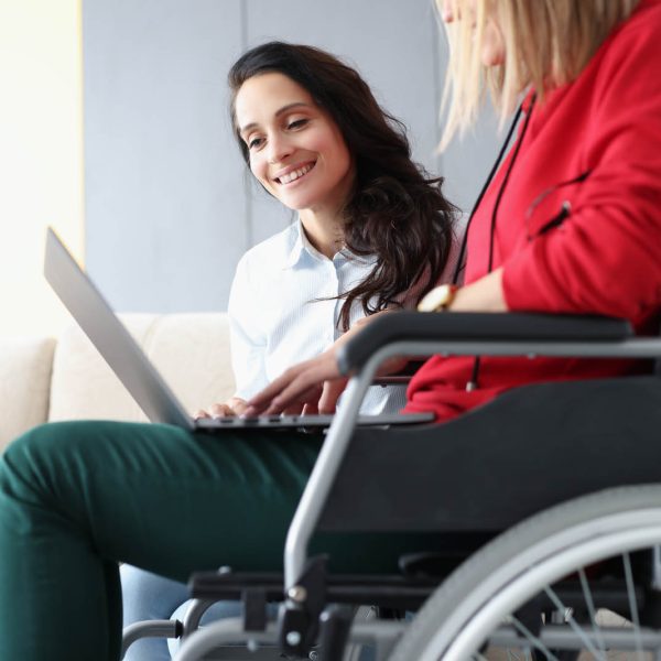 Disabled woman in wheelchair and her friend looking at laptop screen at home Disabled woman in wheelchair and her friend looking at laptop screen at home. Help for people with disabilities concept