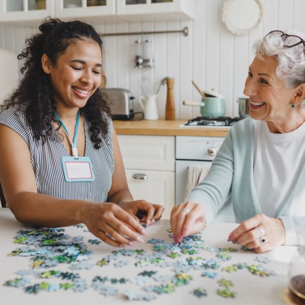 Cheerful senior lady having fun plying puzzle game at kitchen ta Cheerful senior lady having fun plying puzzle game at kitchen table with black female volunteer, taking care of her as social support of retired persons, spending leisure time, talking